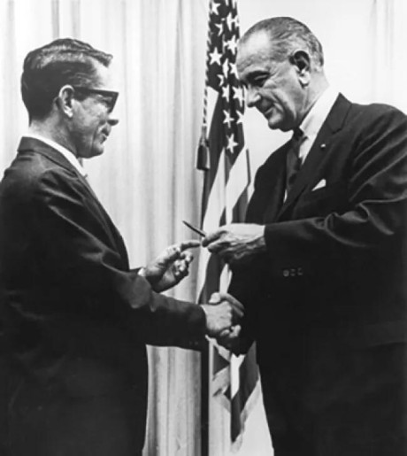 A black-and-white photograph shows two men in suits shaking hands in a formal setting.