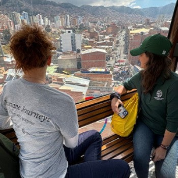 Two people look out a window over a dense population center with mountains in the background.