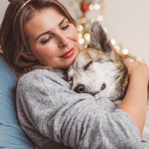 Woman and her dog cuddling on the couch with holiday lights behind them