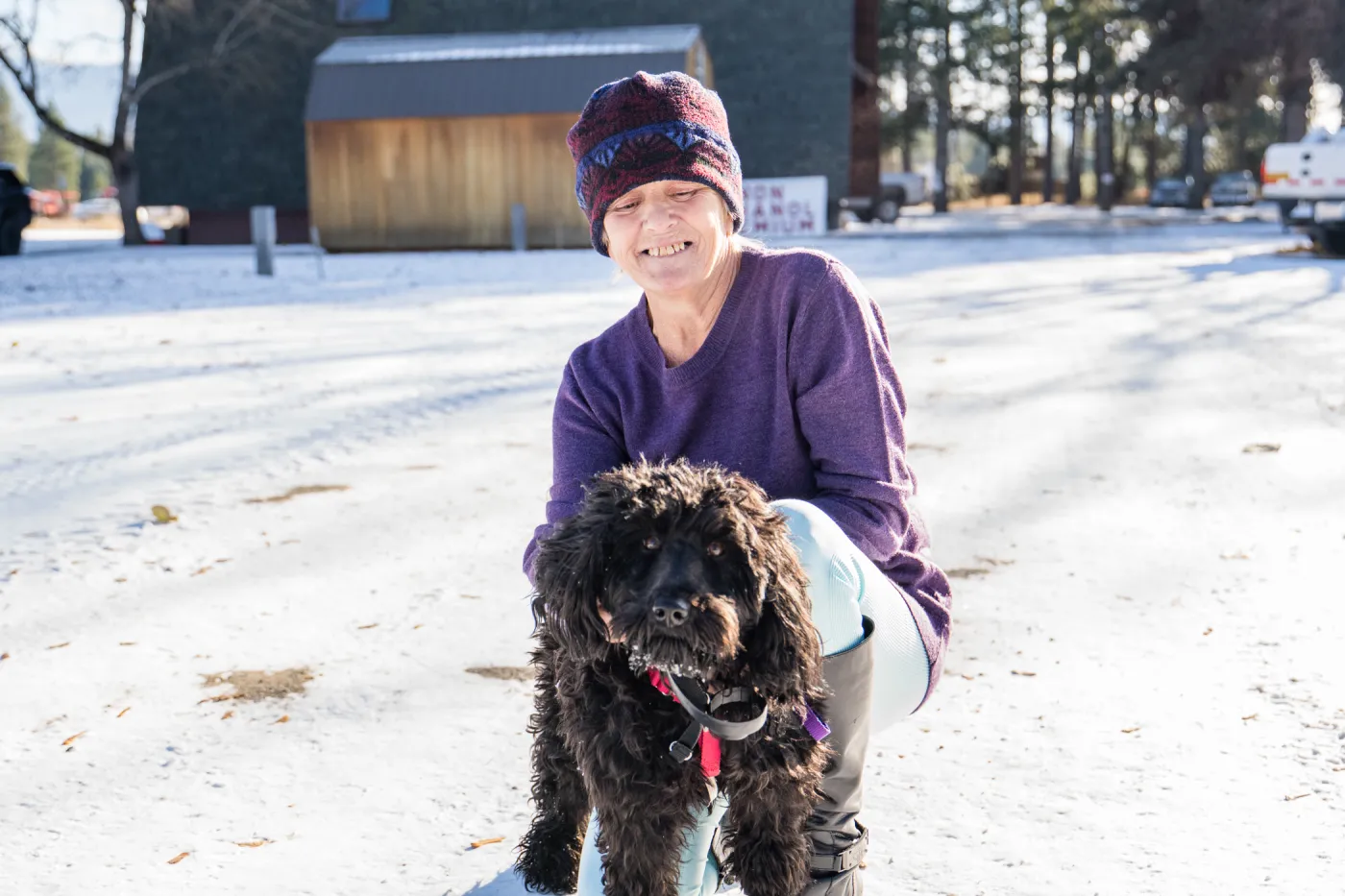 A woman in a purple beanie and shirt holds a black, curly haired dog in the snow