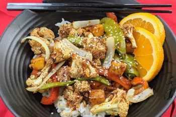 A stir-fry dish with roasted cauliflower, snap peas, bell peppers, and tofu served over white rice, garnished with sesame seeds and orange slices. 