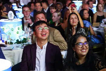 A crowd of people sitting at tables look at a place off screen and smile