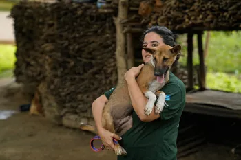 A person smiles while holding a medium-sized brown dog in their arms.
