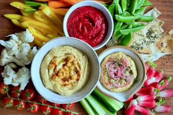 A colorful snack platter is arranged on a wooden board with three dips at the center