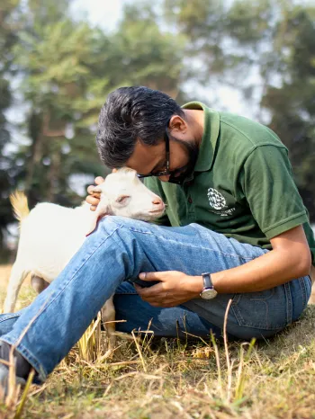 A man in a green shirts sits in a grassy field, embracing a white goat 