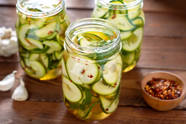 open jars of pickled sliced zucchini and other ingredients for pickling on a wooden table.