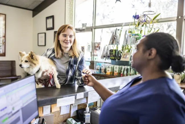 Young woman with small dog paying for veterinary services at veterinarian's office front desk