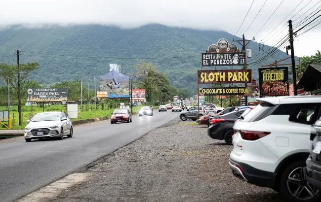 Roadside view of a sloth tourism area with cars, restaurant signs and “Sloth Park” advertising set against forested mountains.