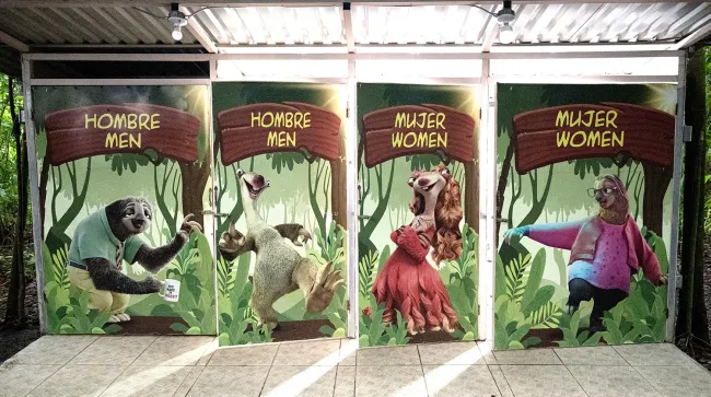 Restroom doors at a sloth park decorated with cartoon sloth characters and signs labeled for men and women in Spanish and English.
