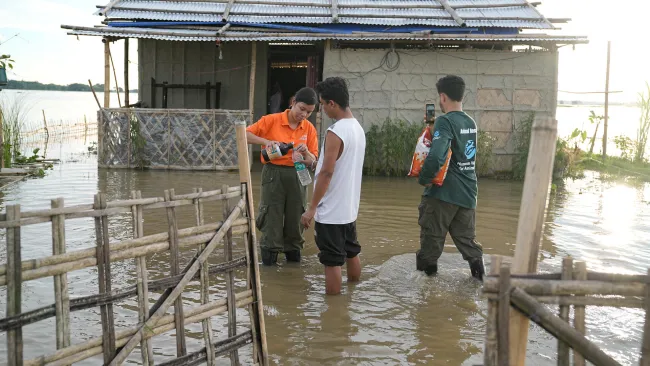 Three people stand in knee-deep floodwater outside a small, raised house