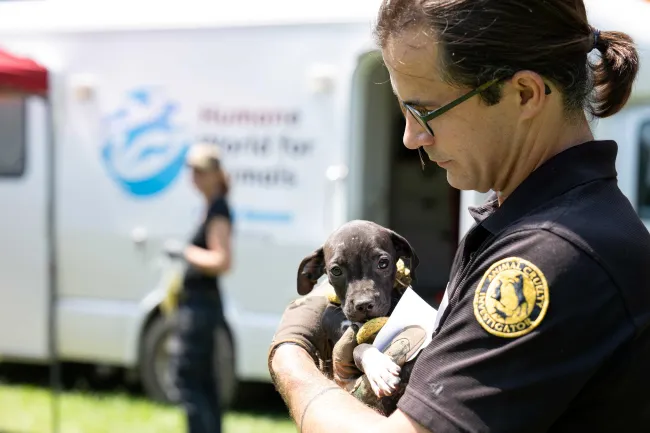 A man wearing holds a small black puppy in his arms. 