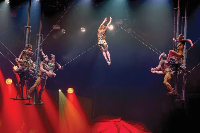 Aerial acrobats performing on a trapeze under colorful stage lights.
