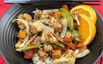 A stir-fry dish with roasted cauliflower, snap peas, bell peppers, and tofu served over white rice, garnished with sesame seeds and orange slices. 
