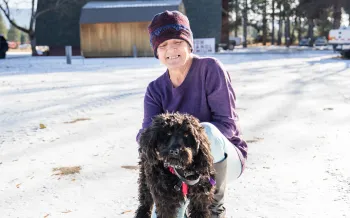 A woman in a purple beanie and shirt holds a black, curly haired dog in the snow
