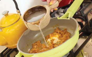 Pouring broth into a skillet of pasta sauce on a stovetop.