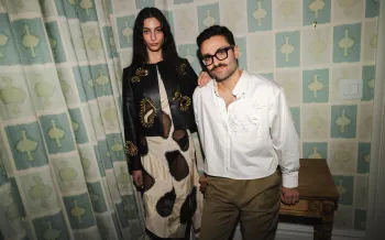 Two people pose indoors against patterned wallpaper.