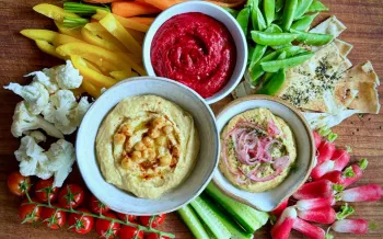 A colorful snack platter is arranged on a wooden board with three dips at the center