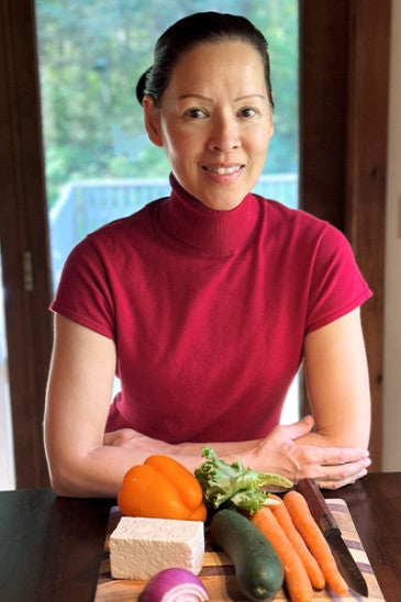 A woman sitting at a table in front of a cutting board with plant-based foods.
