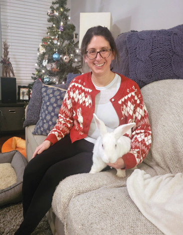 Person sitting on a couch holding a white rabbit beside a Christmas tree.